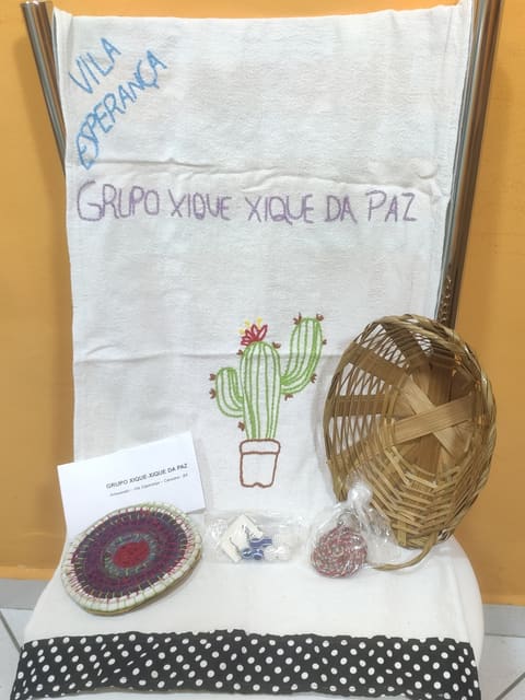 Kit montado sobre uma cadeira: pano bordado com 'Vila Esperança', 'Grupo Xique Xique da Paz' e um cacto, cestinha de palha, descanso de crochê, chaveiro e brinco em saquinhos e um cartão do grupo.