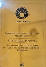 Placa de acrílico com o logotipo da Unicamp e o texto: “Homenageada da 1ª turma do MBE Unicamp. Laura Elisa Alves Amorim. Seu legado persiste na vida de todos os corações que tocou. Parabéns!”