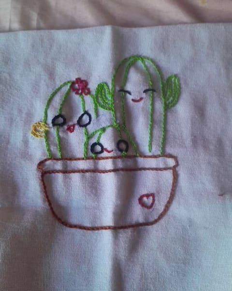 Bordado em andamento num pano claro: três cactos de carinha sorridente num vaso marrom, com uma flor vermelha.
