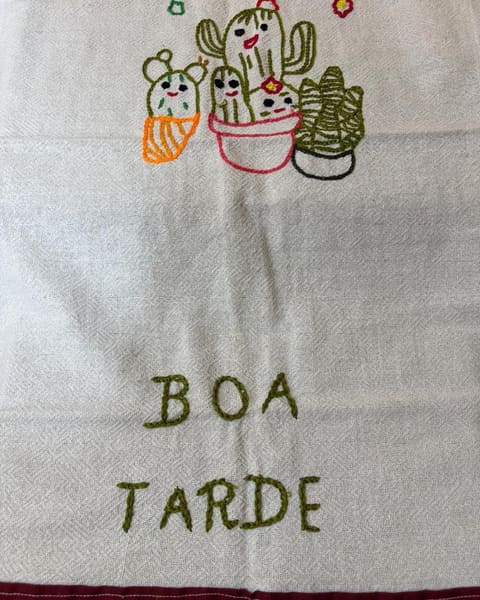 Pano de prato bordado à mão com quatro cactos de vaso, de carinhas sorridentes e florzinhas vermelhas, e as palavras BOA TARDE bordadas em verde. Embaixo, a barra do pano em tecido vinho.